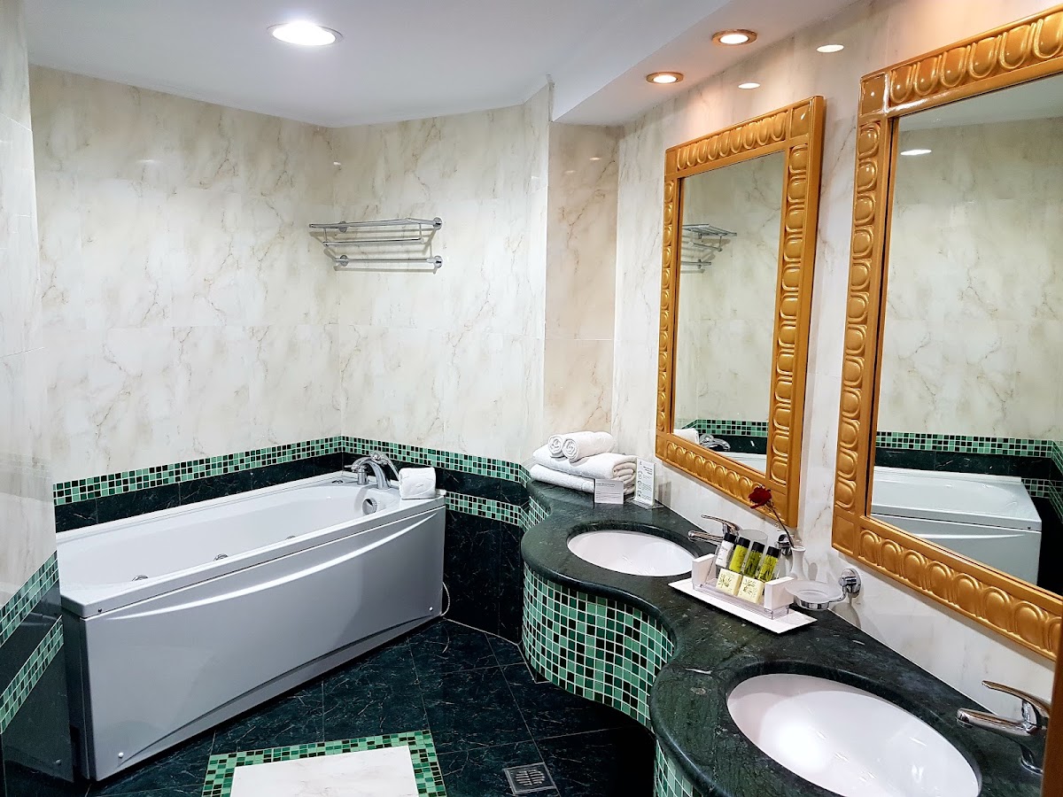 Bathroom with marble walls, curved soaking tub, dual sinks with green mosaic trim, gold-framed mirrors, and white towels on counter.