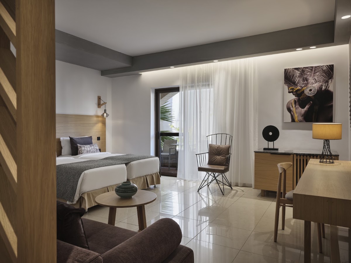 Modern resort suite with queen bed, sitting area, desk, and balcony access overlooking tropical grounds.