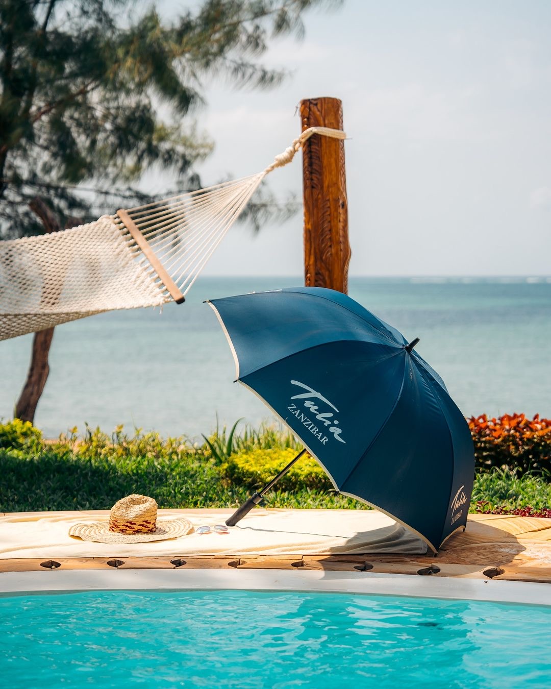 Beachside hammock and umbrella at Tulia Zanzibar with ocean view, straw hat, and pool edge in foreground.