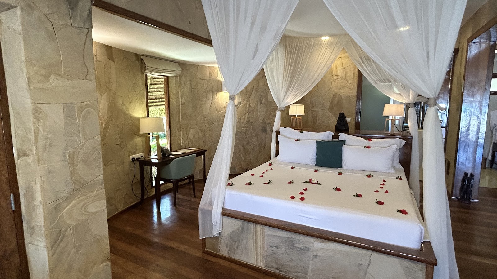Four-poster canopy bed with white drapes and rose petals, stone walls, desk area, and warm lighting in a resort suite.