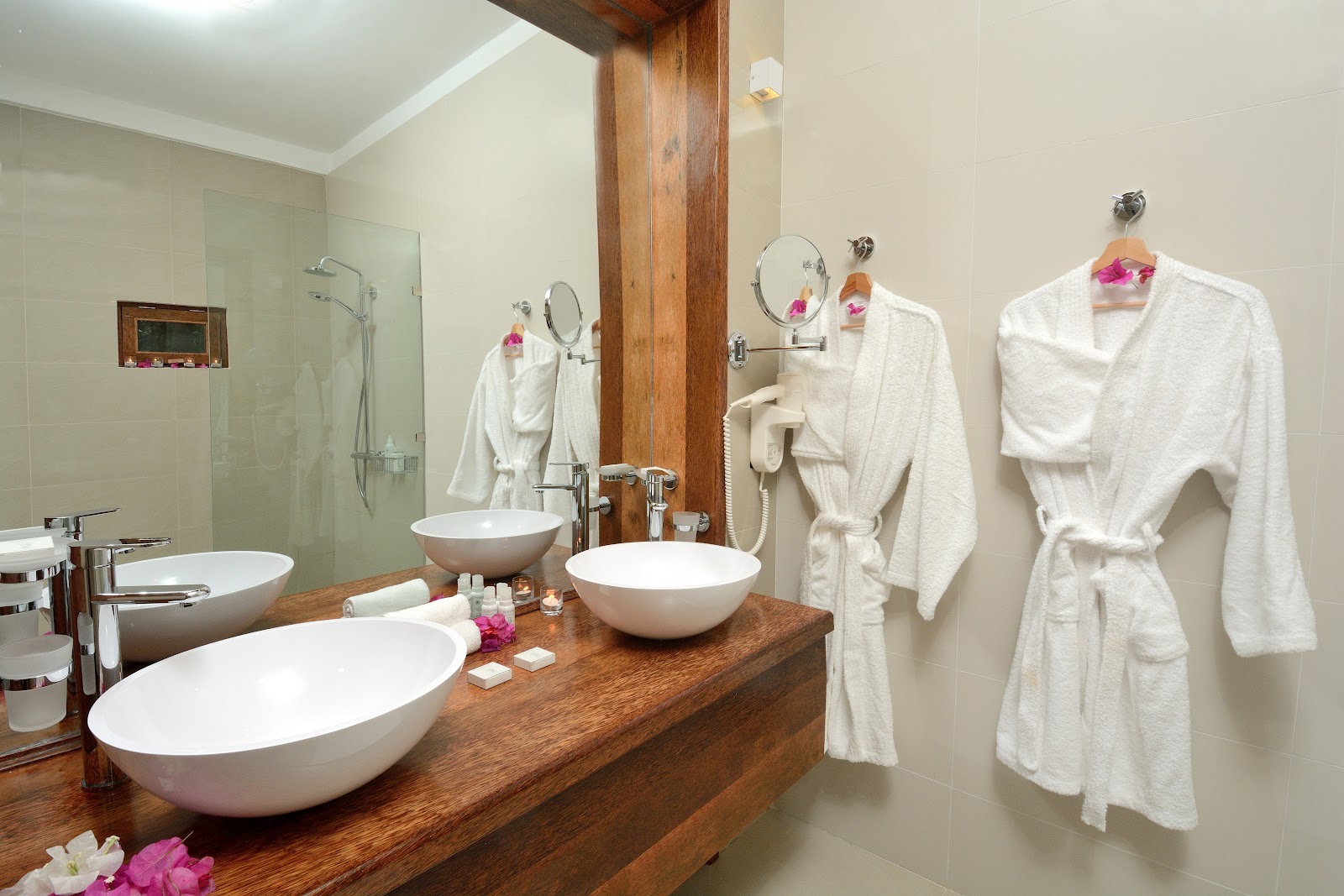 Spa bathroom with wooden vanity holding three white vessel sinks, fluffy white robes on wall hooks, and pink flowers as décor accents.