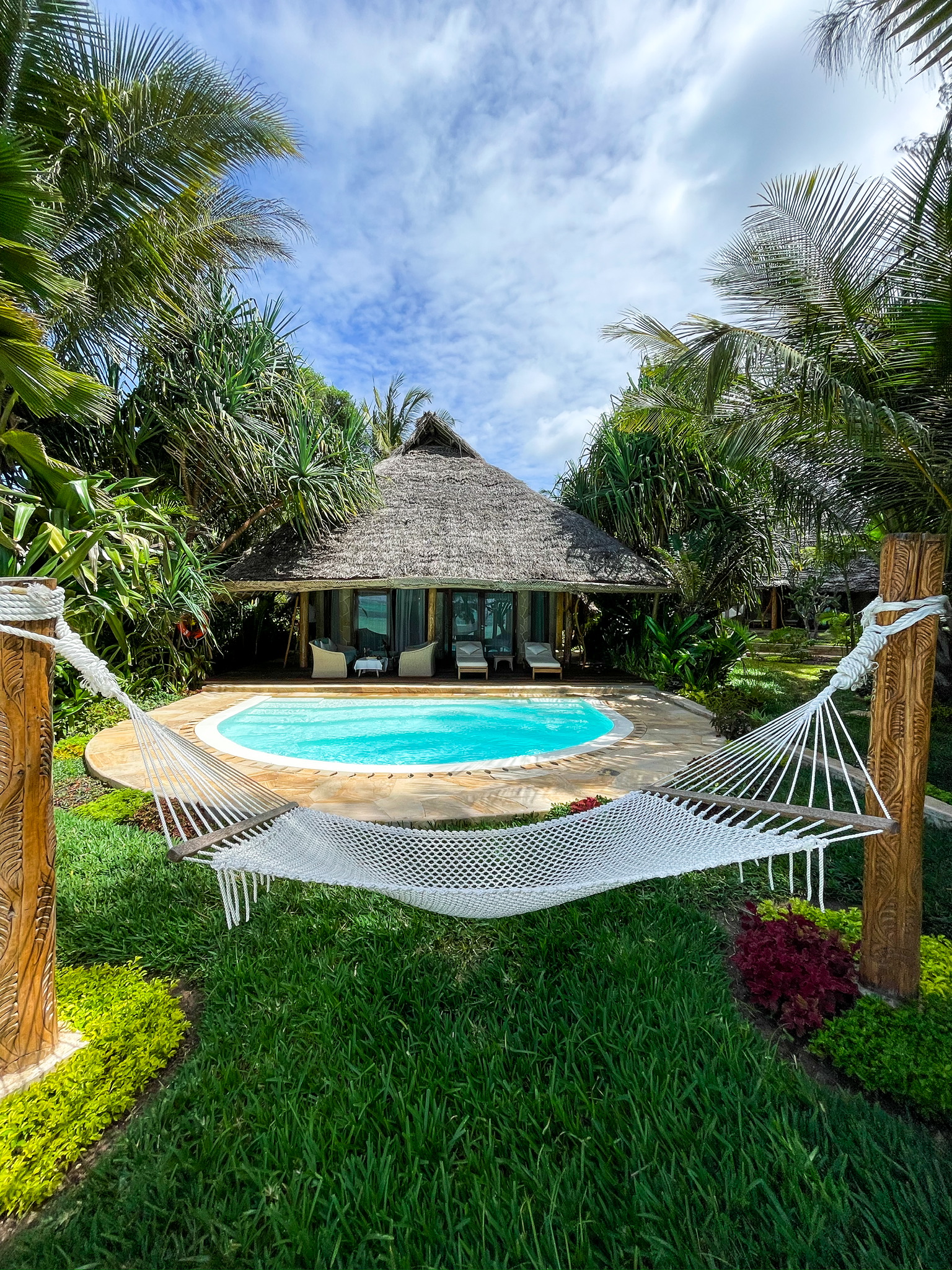 Thatched-roof bungalow with wooden deck and lounge chairs overlooks white sand beach and turquoise water at Tulia Zanzibar, with palm fronds framing the scene.