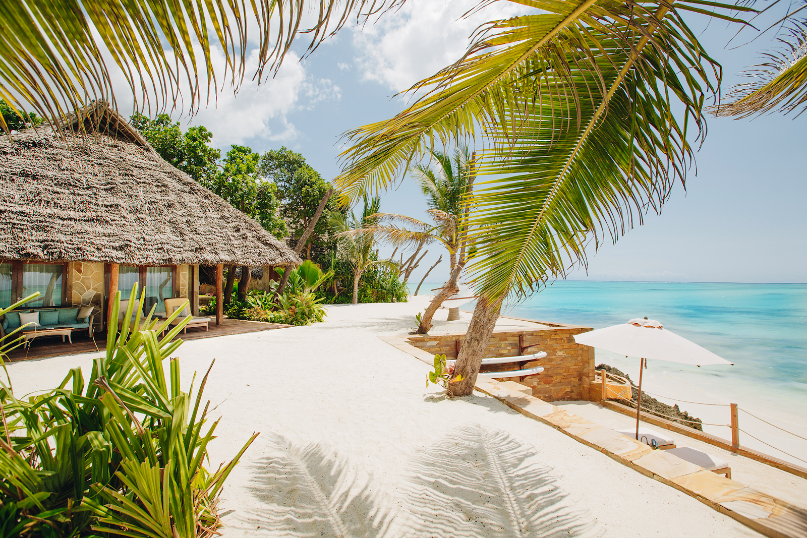 Turquoise pool surrounded by wooden decking at Tulia Zanzibar Unique Beach Resort, with thatched roof structures and lush tropical gardens beyond.