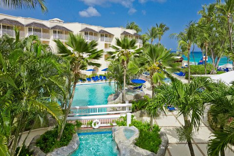 White multi-story resort buildings overlook a turquoise pool surrounded by palm trees and blue lounge chairs on a sunny day at Turtle Beach, Barbados.