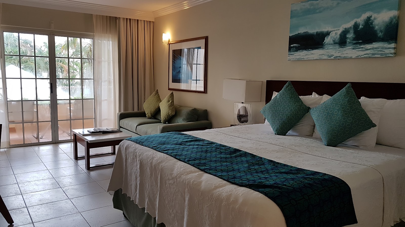 Resort guest room with king bed, teal accent pillows, seating area, and balcony overlooking palm trees at Turtle Beach Barbados.