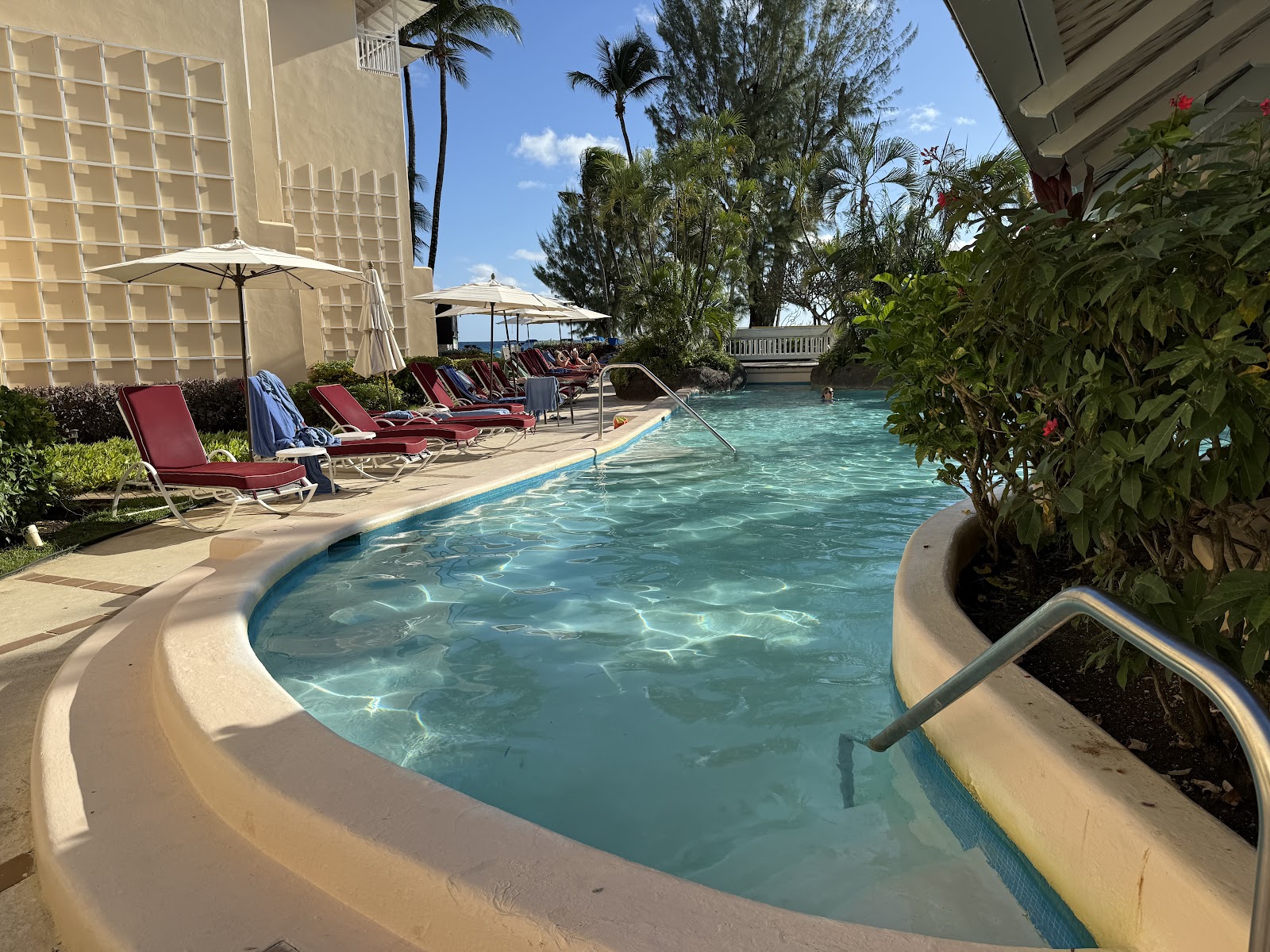 Curved pool with turquoise water beside red lounge chairs and umbrellas at Turtle Beach resort, with cream-colored buildings and palm trees in background.