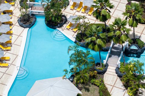 Overhead view of a bright blue pool at Turtle Beach Barbados with palm trees, yellow lounge chairs, and white umbrellas on the surrounding deck.
