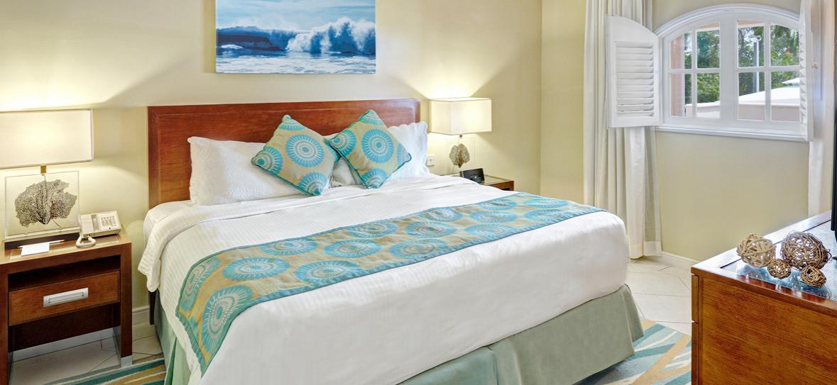 Resort bedroom with wood headboard, white linens, turquoise patterned pillows and runner, cream walls, and ocean artwork above the bed.