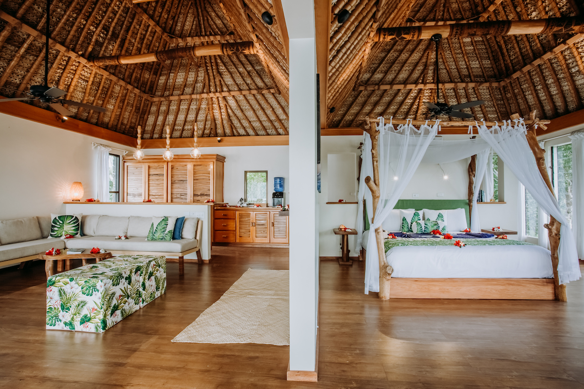 Open-plan bungalow interior with wooden beamed ceilings, woven thatch roof, cream sofa, four-poster bed with white curtains, and tropical leaf decor.