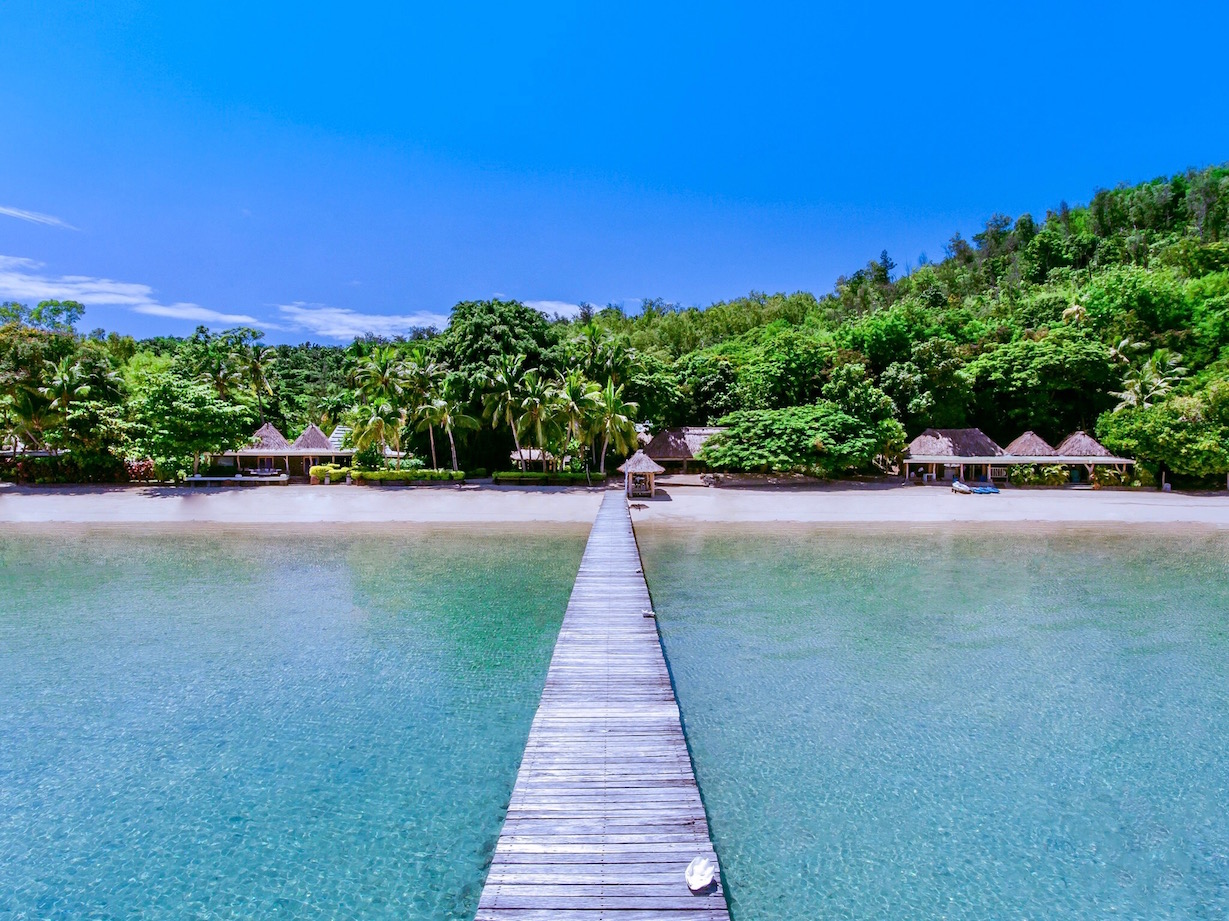 Wooden jetty extending into turquoise water toward a sandy beach lined with thatched bungalows and lush tropical forest at Turtle Island Resort Fiji.