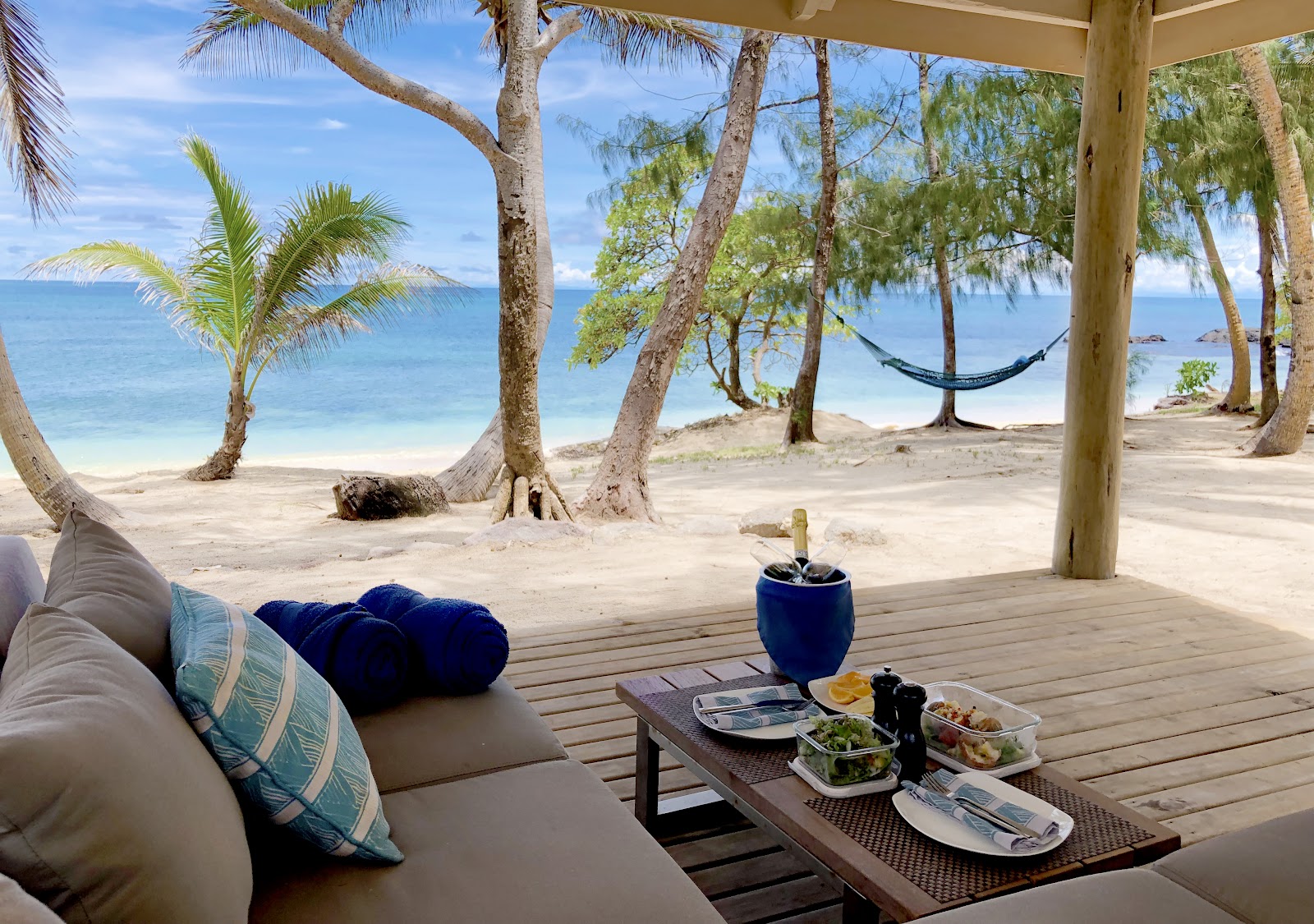 Covered deck with cushioned seating, meals, and ocean views framed by palm and casuarina trees, with a hammock and sandy beach beyond.