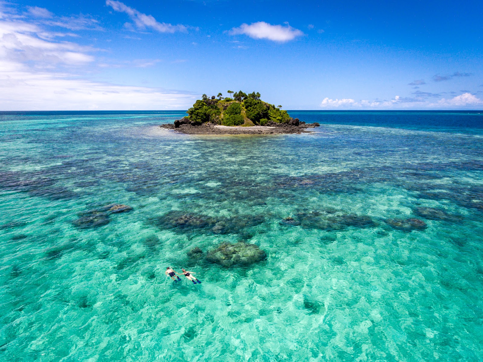 Two snorkelers in clear turquoise water with a small forested island and coral reefs visible in the distance.
