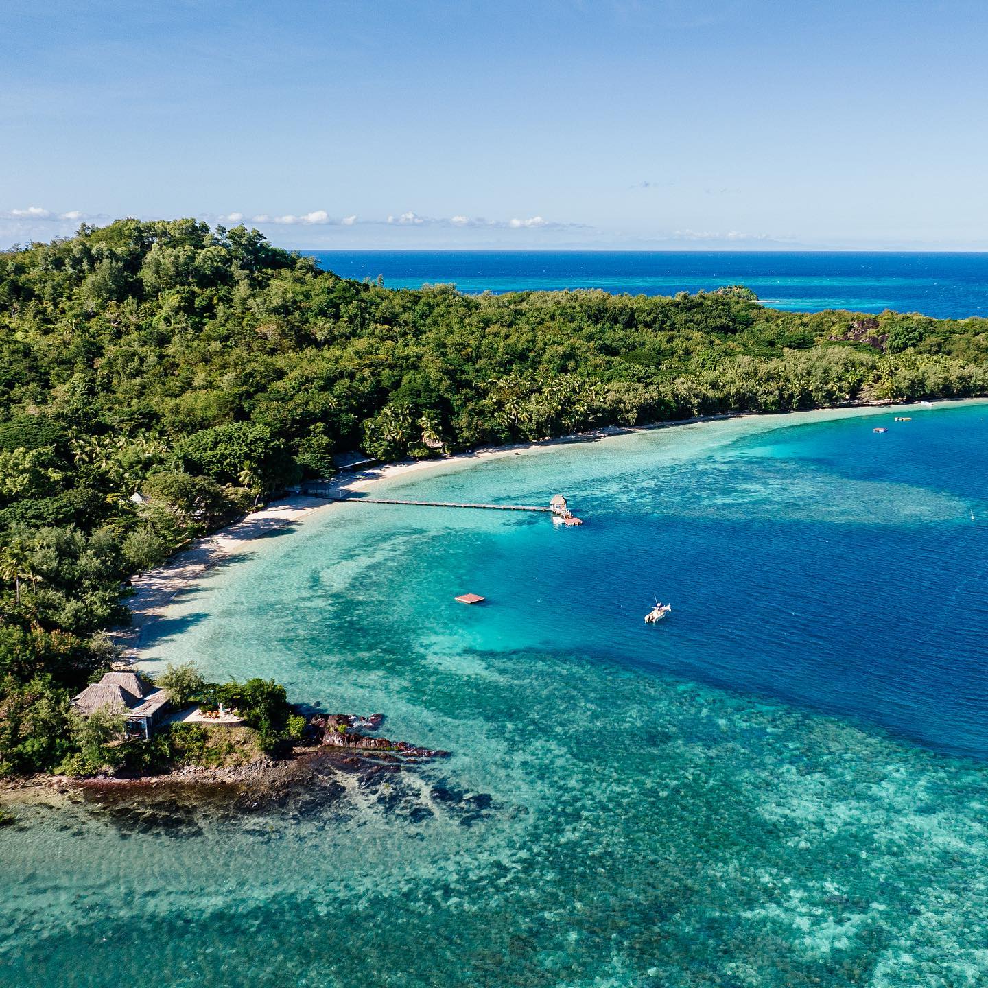 Aerial view of Turtle Island Resort Fiji's turquoise lagoon with sandy beach, dense jungle, moored boats, and a thatched-roof structure on the shore.