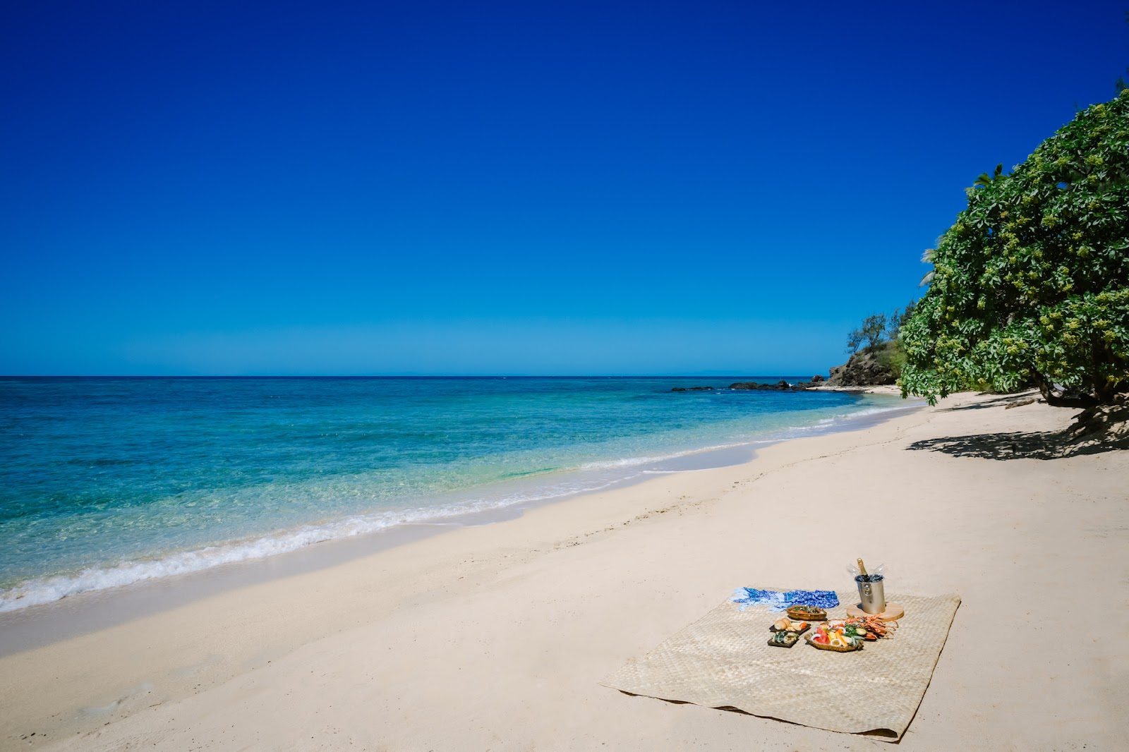 Picnic setup on white sand beach at Turtle Island Resort Fiji with turquoise water and tropical vegetation beyond.