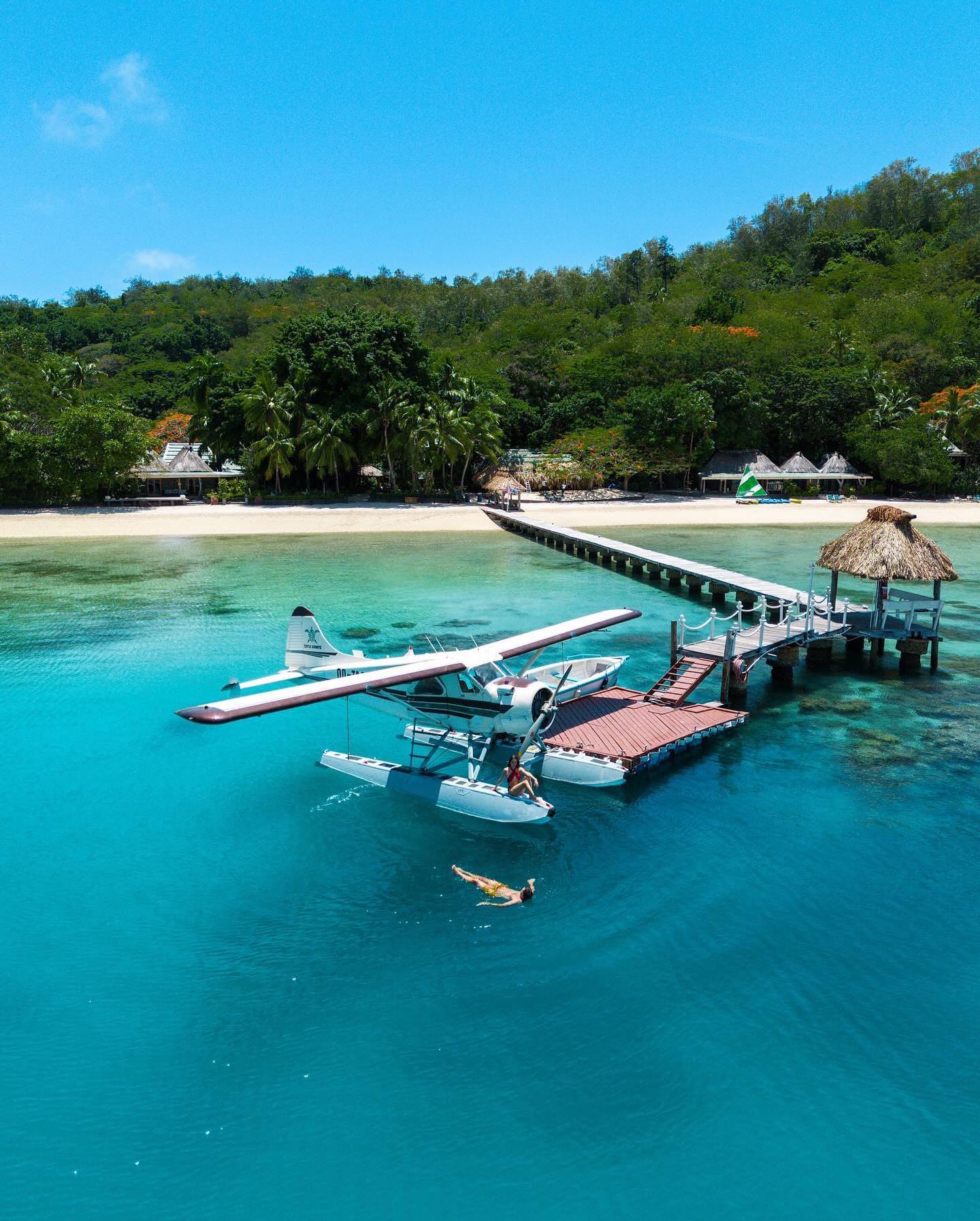 Seaplane moored at a wooden dock with thatched-roof structures, a snorkeler in turquoise water, white sand beach, and forested hillside at Turtle Island Resort Fiji.