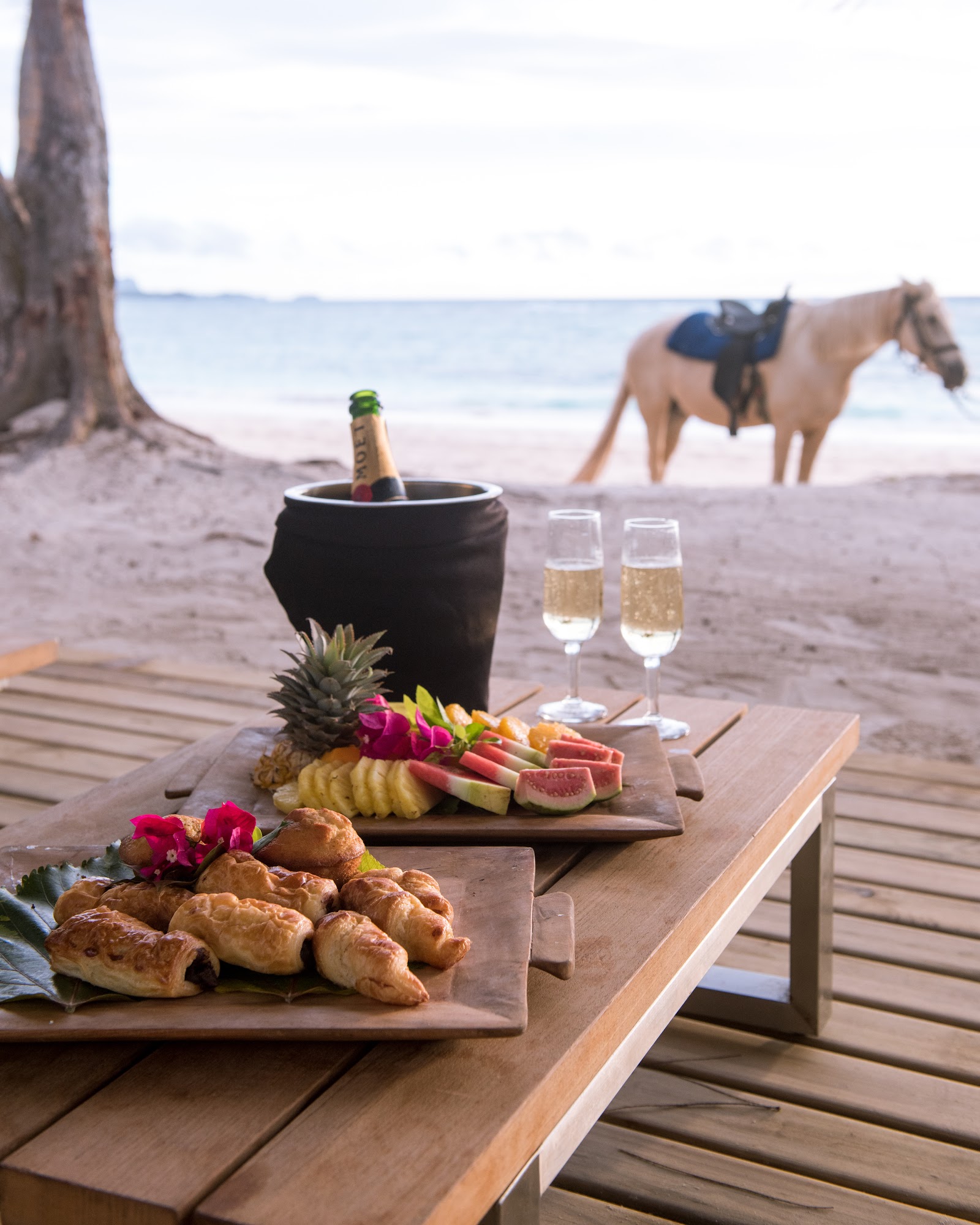Beach table set with champagne, pastries, and tropical fruit at Turtle Island Resort Fiji, with a horse and rider in the sand beyond.