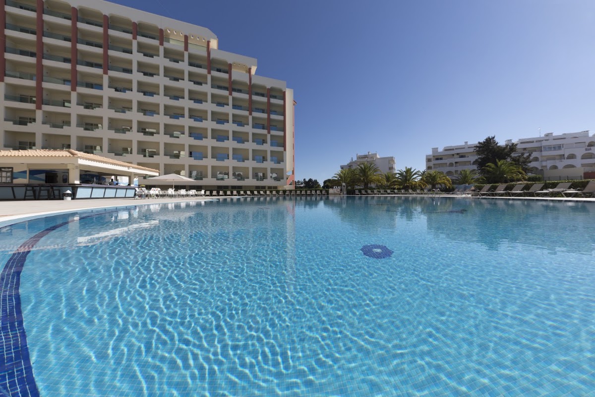 Large resort pool with clear blue water, lounge chairs, and multi-story hotel buildings with red trim under clear sky.
