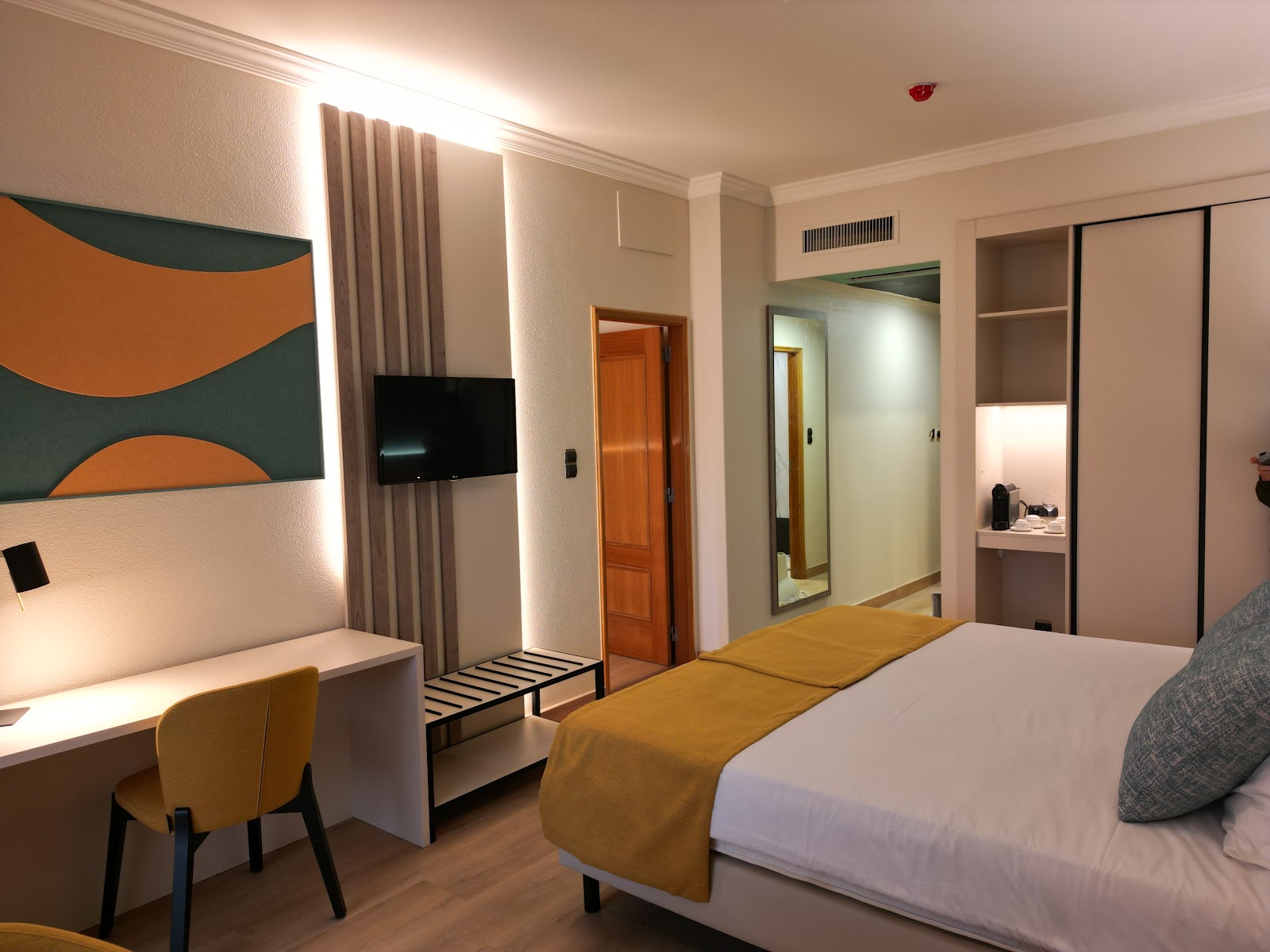 Modern resort bedroom with queen bed, blue pillows and throw, wood headboard with table lamps, cream armchair, and balcony overlooking another building.