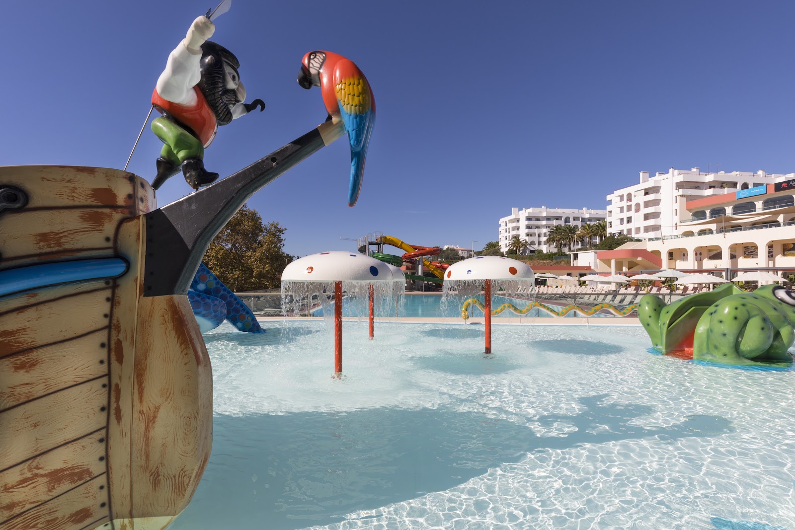 Pirate-themed water park with animatronic figures, shallow pool, and white resort buildings under clear blue sky.