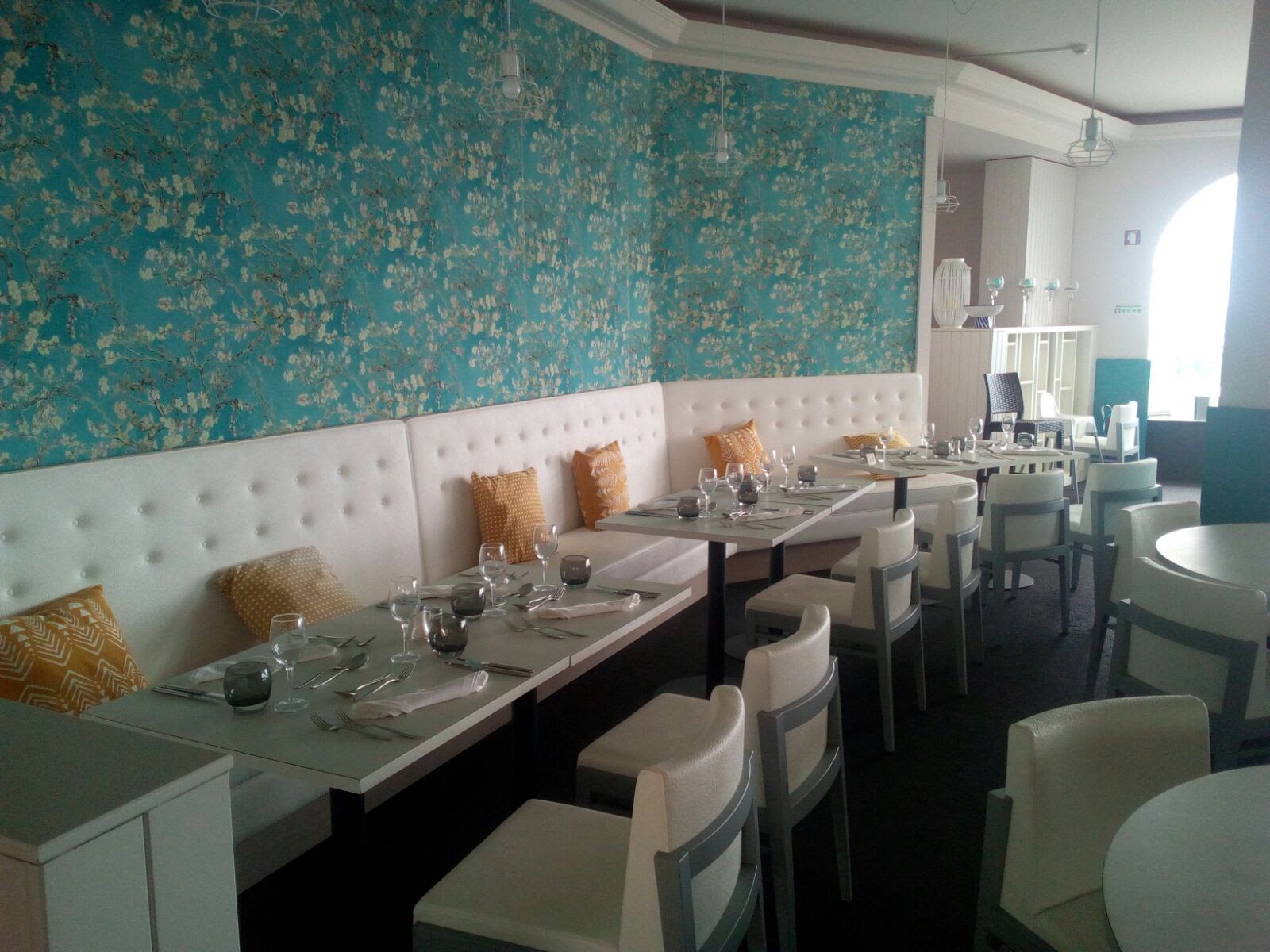 Restaurant dining room with turquoise floral wallpaper, white banquette seating, and tables set with place settings and glassware.