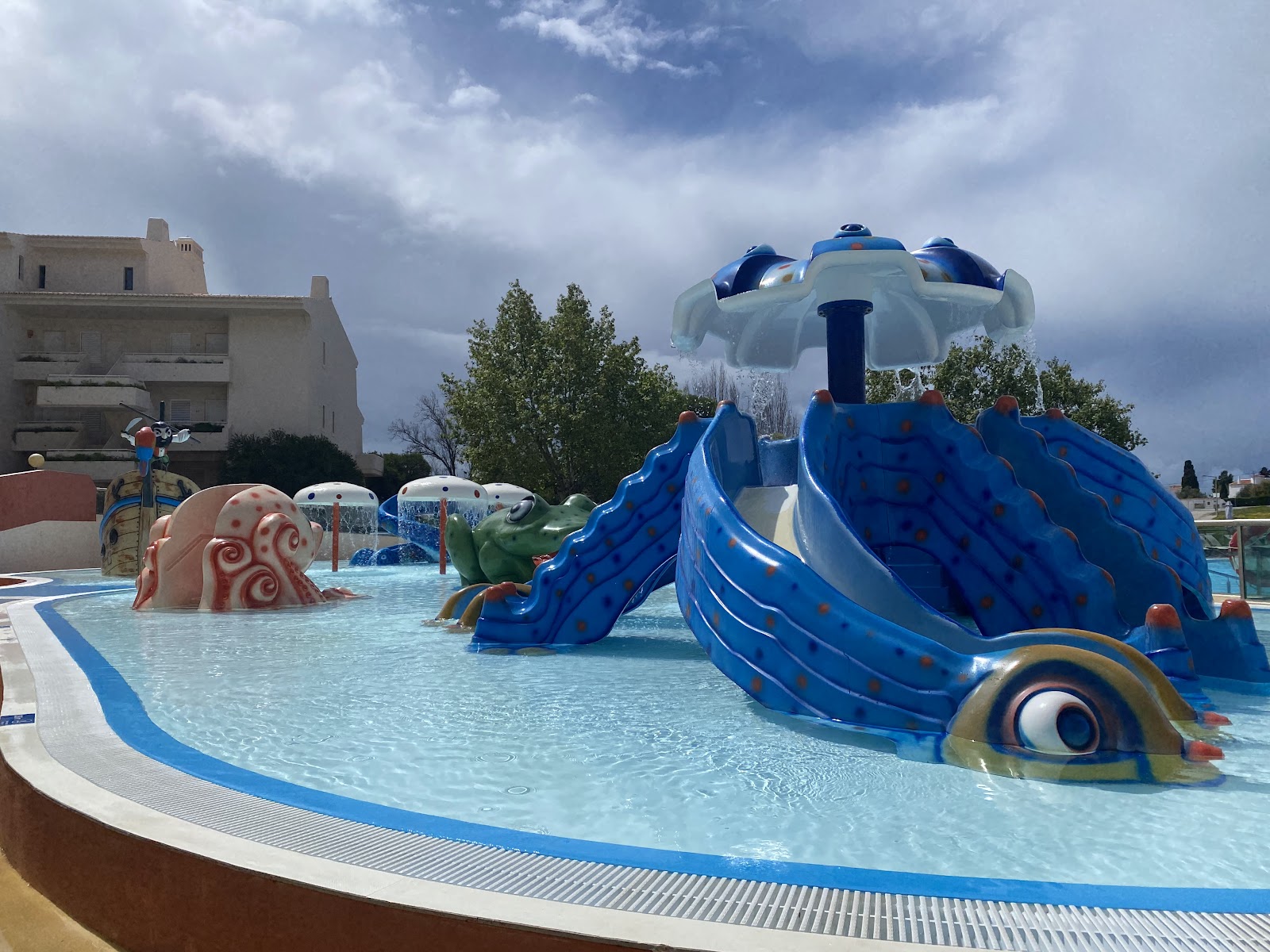 Multi-story resort buildings with red and gold accents surround a large blue pool and sprawling green lawn dotted with lounge chairs and umbrellas under clear skies.