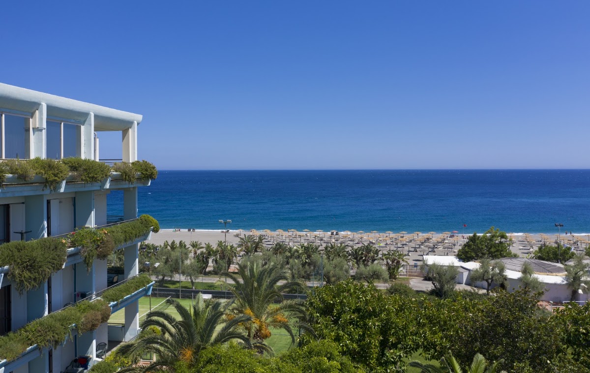 Modern white resort building overlooking a beach lined with umbrellas and palm trees, with clear blue sea beyond.