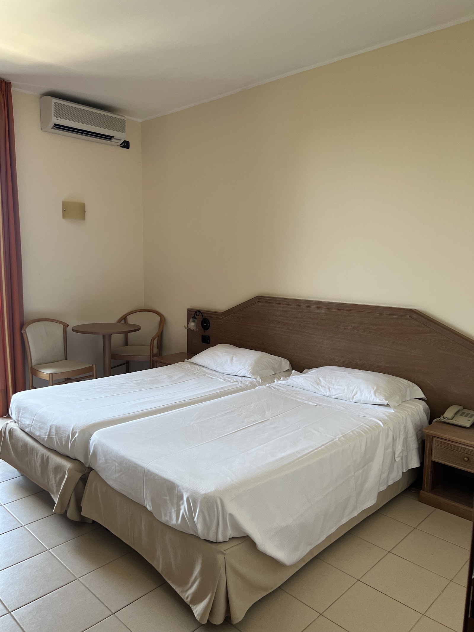 Resort bedroom with two double beds with white linens, wooden headboard, small table and chair, air conditioning unit, and beige walls.