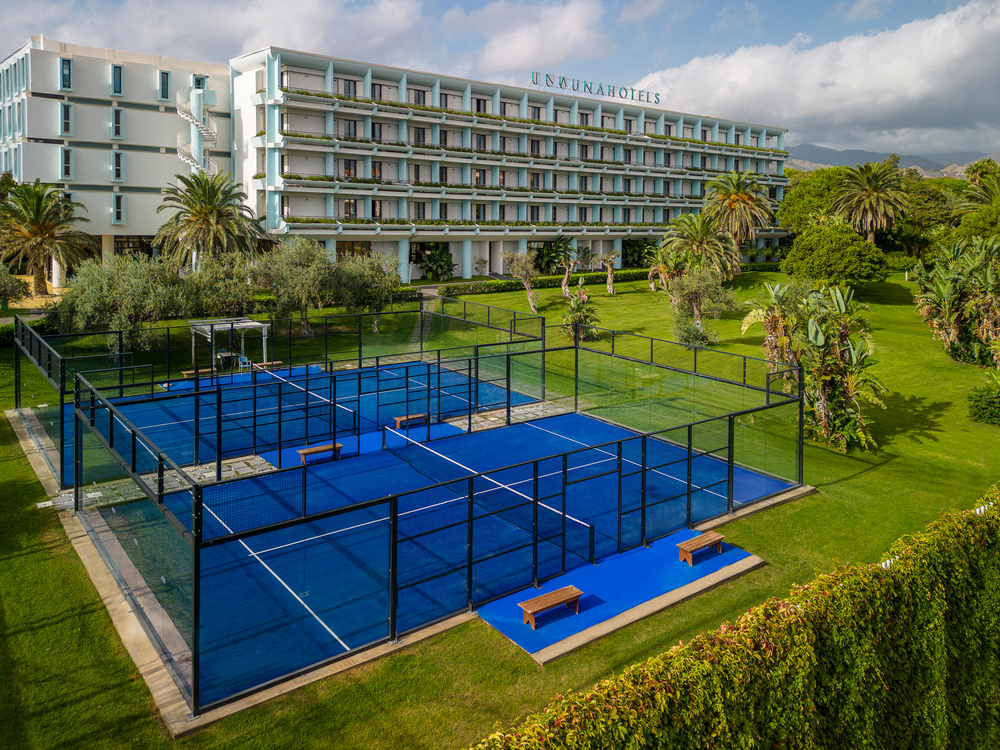 UNA Hotels Naxos Beach Sicilia's blue padel courts on manicured grounds with palm trees and the white resort building in the background.