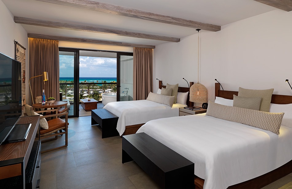 Resort bedroom with two queen beds, wooden beams, ocean view through glass doors, and tropical coastline visible beyond the balcony.