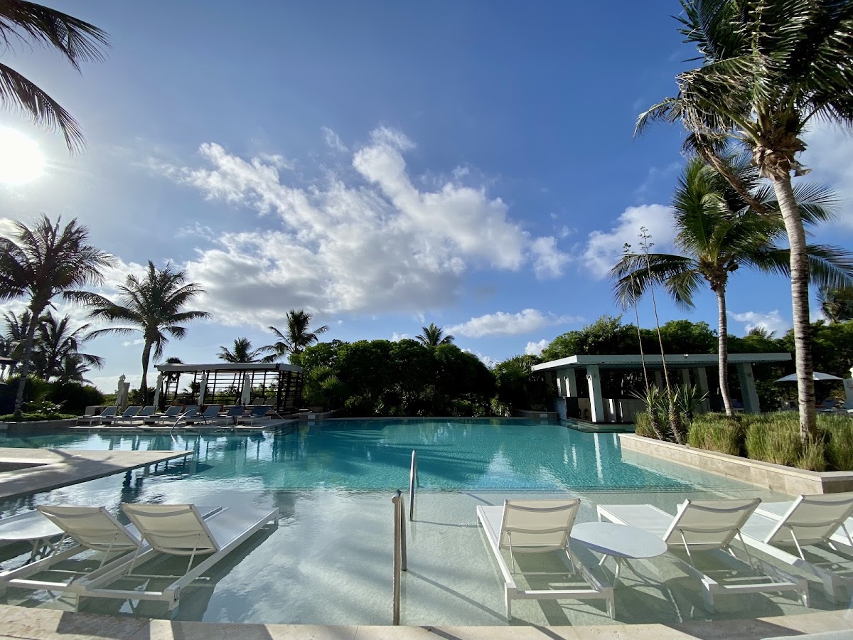 White lounge chairs line the shallow edge of a turquoise pool at UNICO 20°87° Hotel Riviera Maya, with palm trees and a poolside bar visible under blue sky.