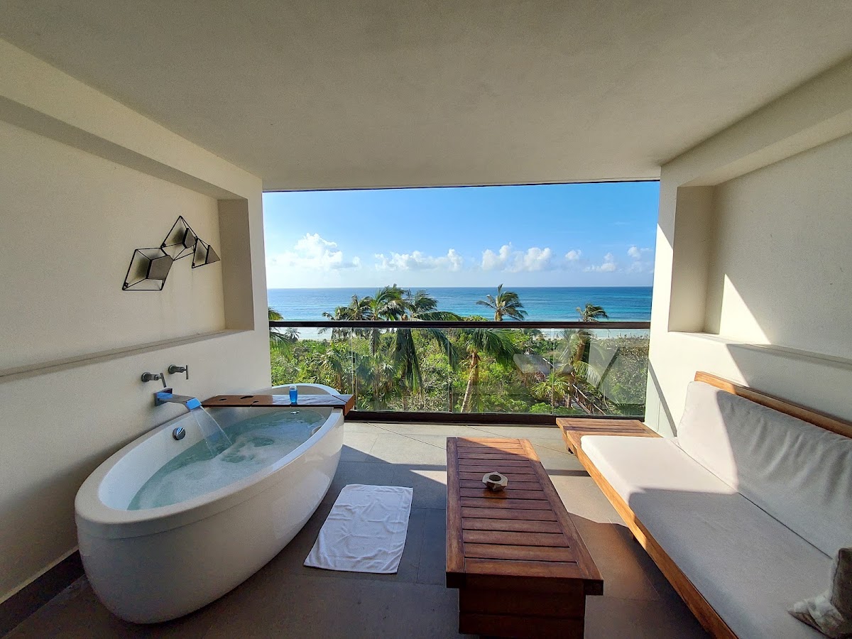 Resort suite bathroom with soaking tub, lounge chairs, and ocean view beyond the balcony with palm trees and turquoise Caribbean water.