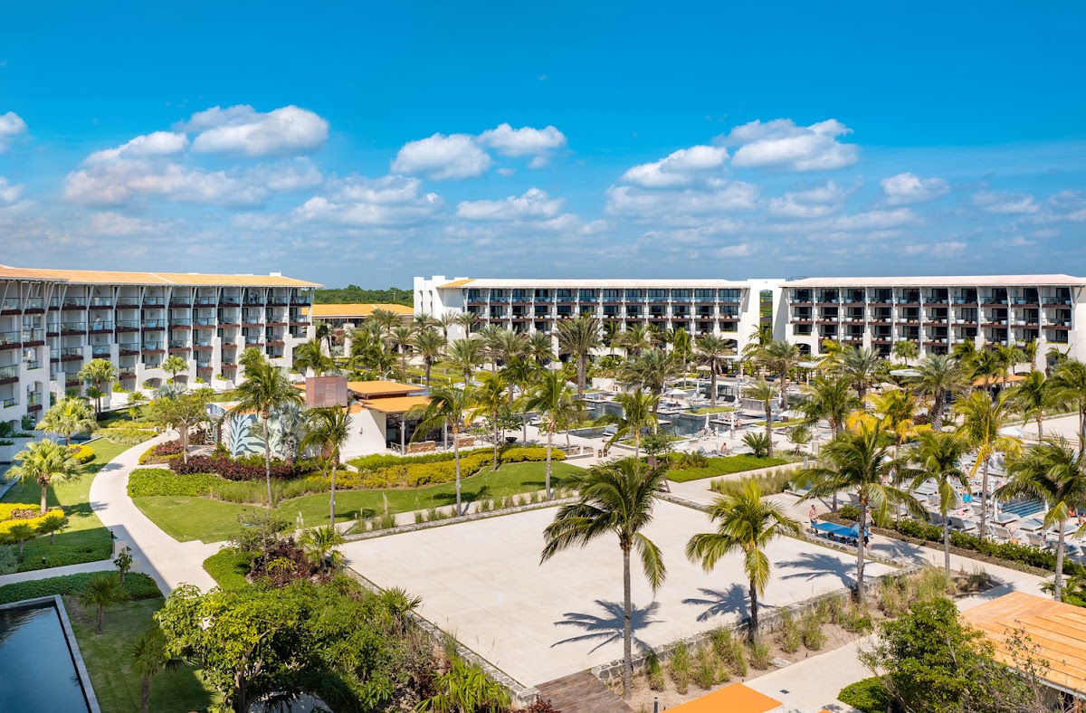 Modern white resort buildings with gold-trimmed roofs overlook manicured grounds, palm trees, and a central courtyard under blue sky with white clouds.