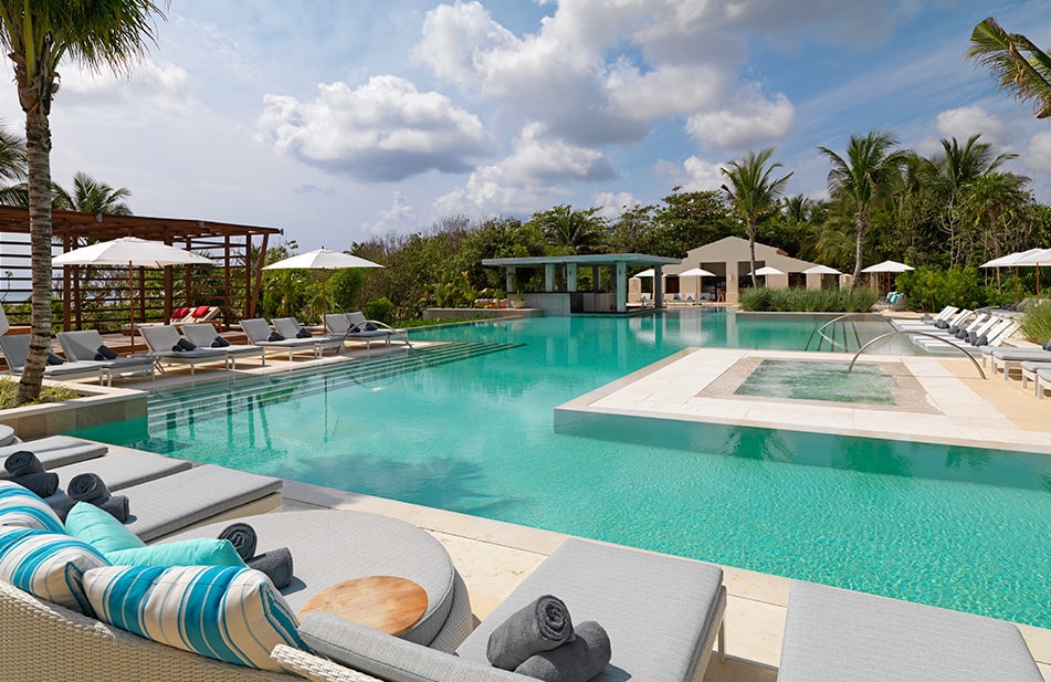 Turquoise pool with lounge chairs, umbrellas, and palm trees surrounding modern resort buildings under cloudy sky.