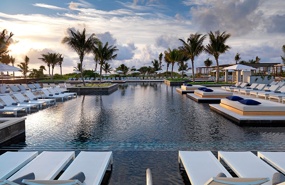 Aerial view of UNICO 20°87° Hotel Riviera Maya with white sand beach, turquoise ocean, blue pool complex, and modern hotel buildings surrounded by tropical vegetation.