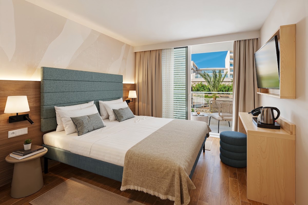 Modern hotel bedroom with gray upholstered bed, balcony overlooking palm trees and coastal buildings under blue sky.