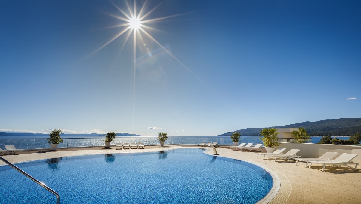 Curved swimming pool and sun loungers on a terrace overlooking a blue bay, with mountains across the water under bright sunshine.