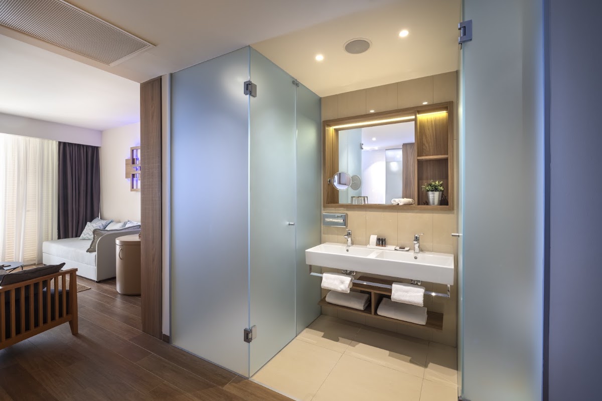 Modern bathroom with double sink vanity, large mirror, and glass-enclosed shower adjoining a resort room with bed and window.