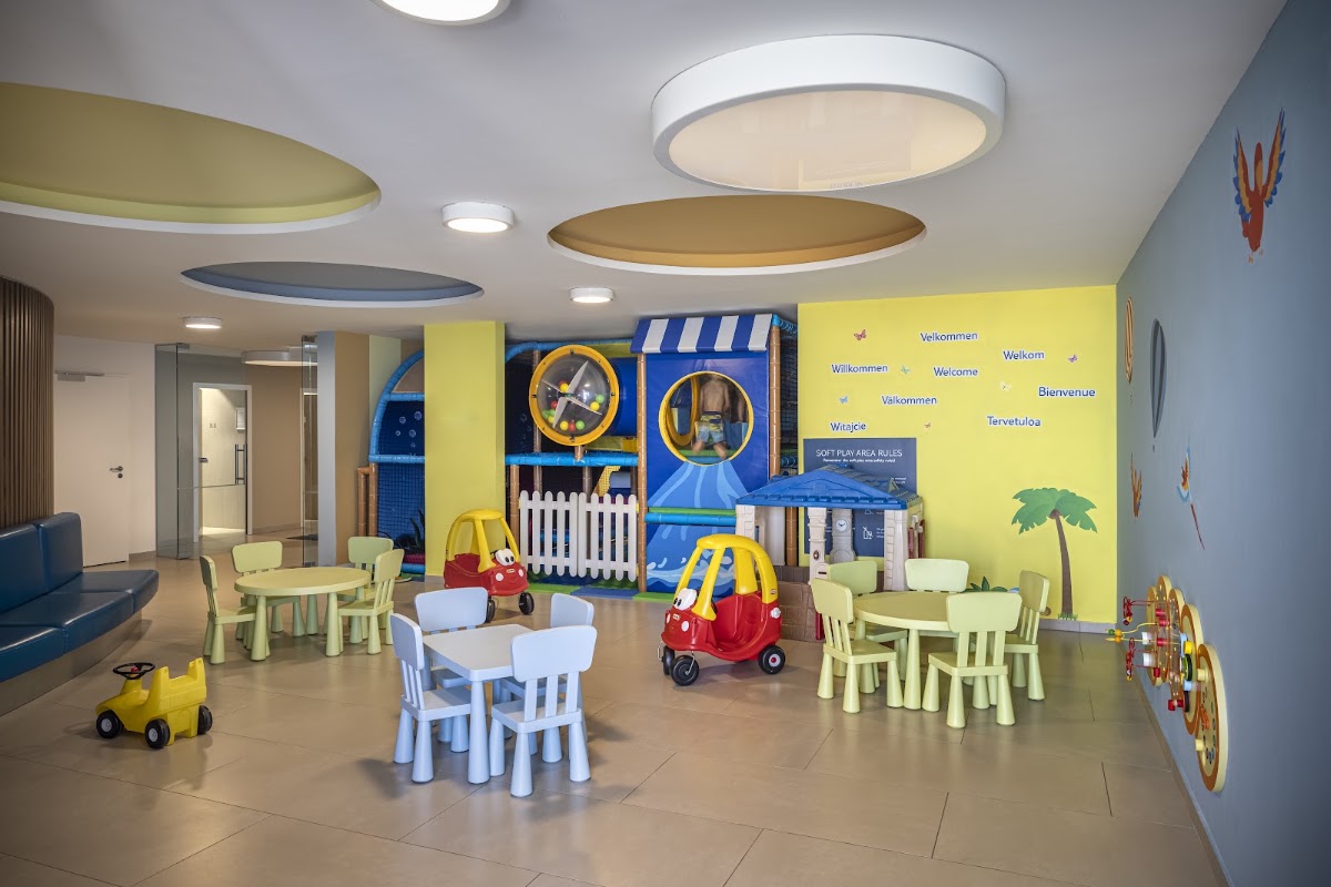 Bright children's play room with small tables and chairs, toy vehicles, play structures, and colorful wall graphics in blue and yellow tones.