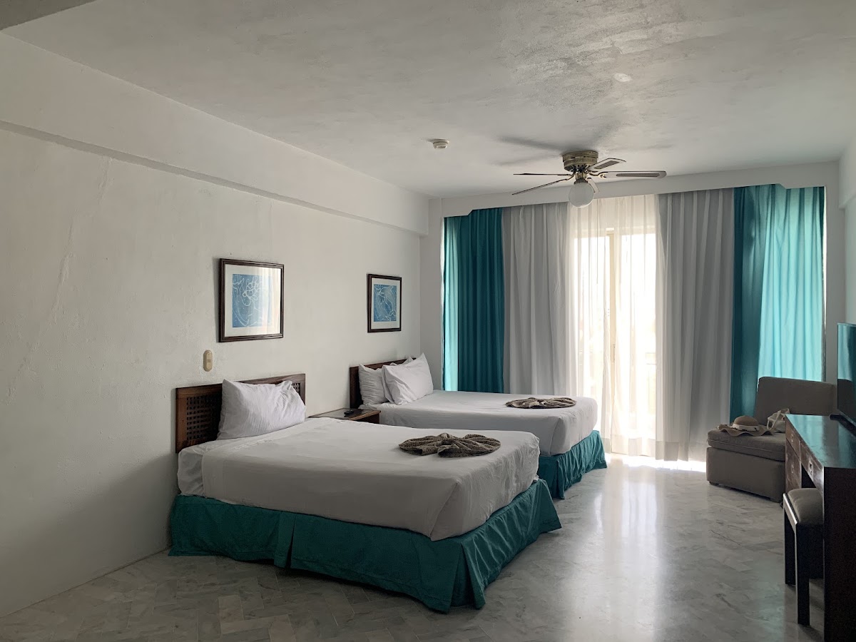 Double beds with white sheets and teal skirts in a resort room with balcony access and ocean-view curtains.