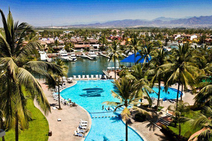 Vamar Vallarta resort's turquoise pool and marina with palm trees, boats, and mountains in the background under clear blue sky.