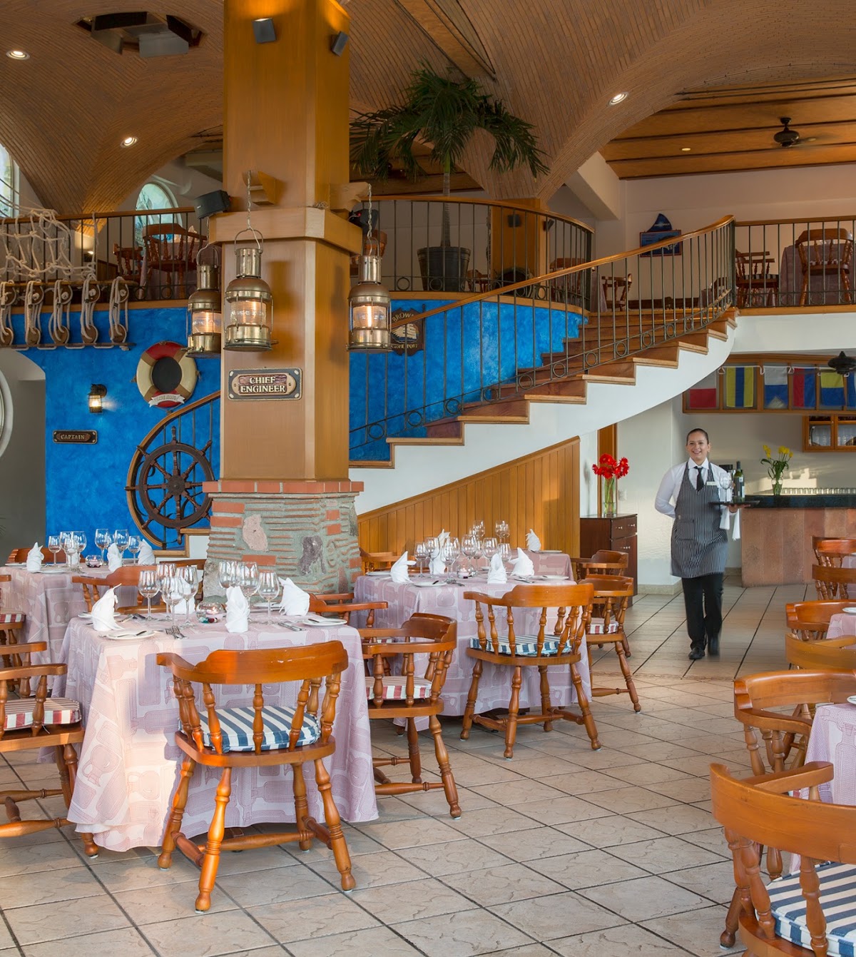 Dining room with nautical theme, wooden chairs around pink-clothed tables, ship wheel and blue wall panels, server in apron standing by bar area.
