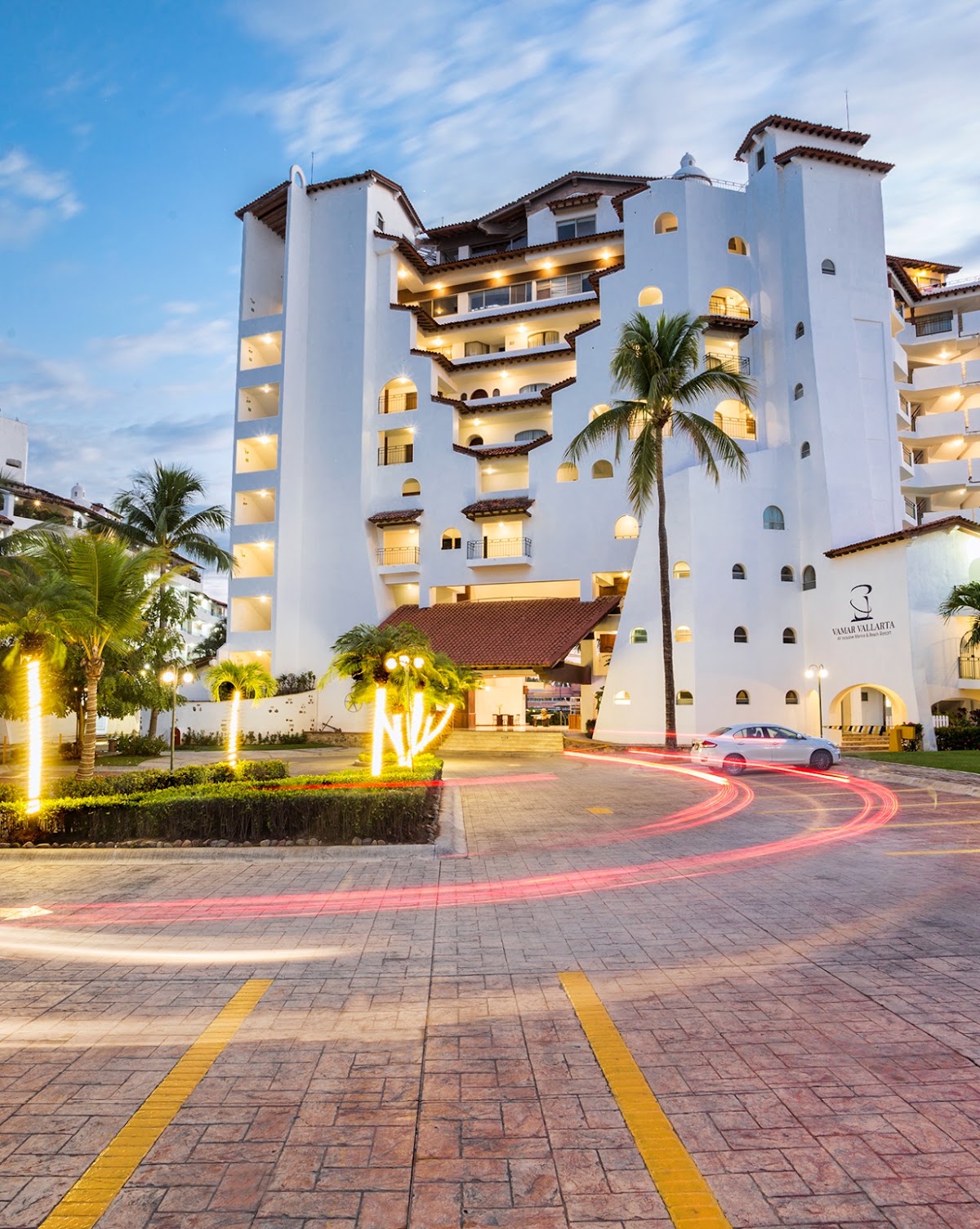 White multi-story resort building with lit windows at dusk, palm trees, and illuminated entrance courtyard with parked car.