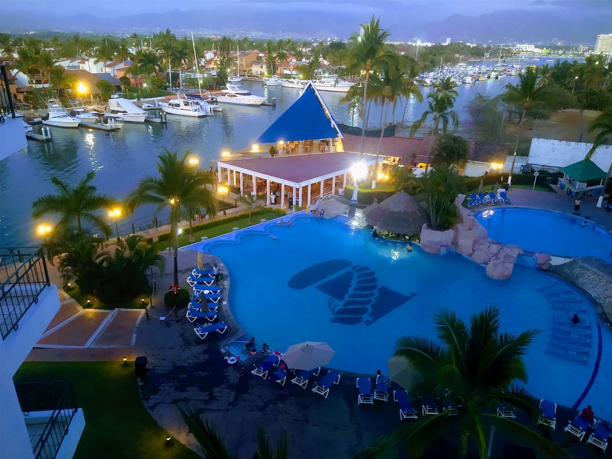 Aerial view at dusk of Vamar Vallarta's blue-roofed pavilion, illuminated pool with lounge chairs, palm trees, and marina filled with boats in the background.