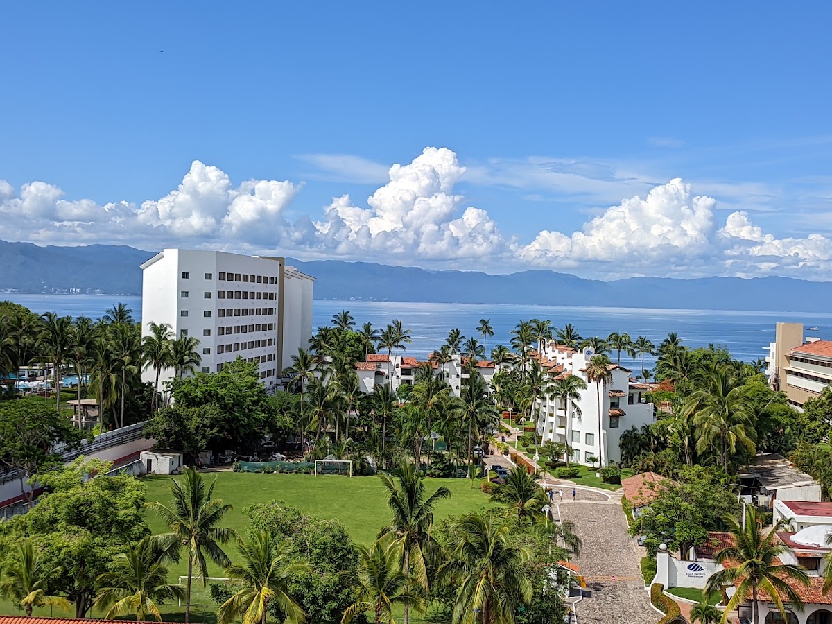 Vamar Vallarta All Inclusive overlooks manicured lawns, palm trees, and the blue Pacific Ocean with mountains in the distance under a cloudy sky.