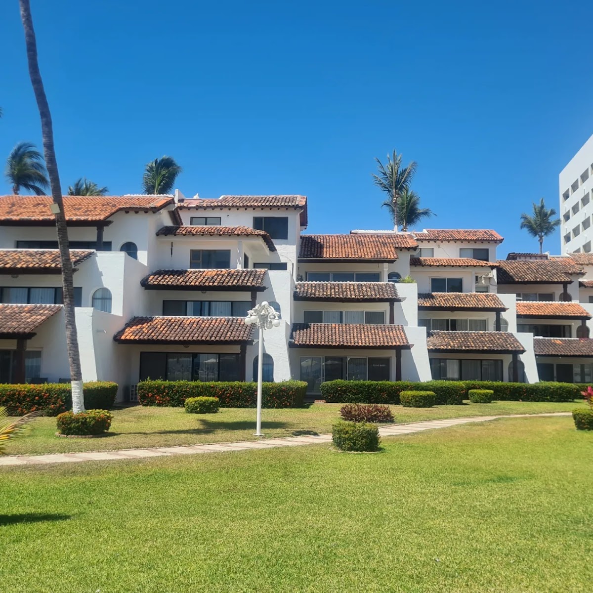 White stucco resort buildings with terracotta tile roofs, palm trees, and manicured lawn under clear blue sky.