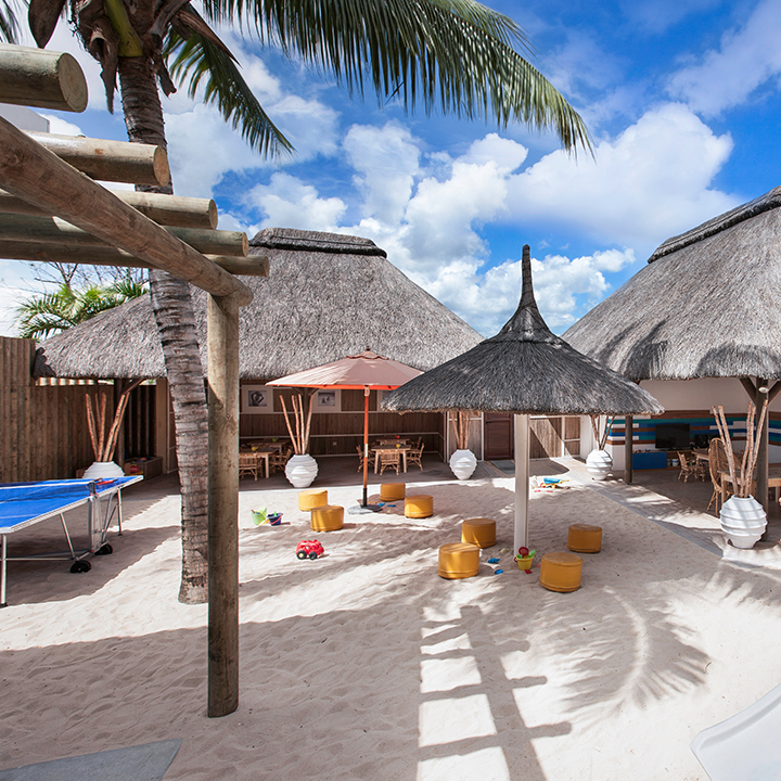 Sandy courtyard at Veranda Pointe aux Biches with thatched-roof bungalows, palm trees, and yellow poufs under umbrellas.