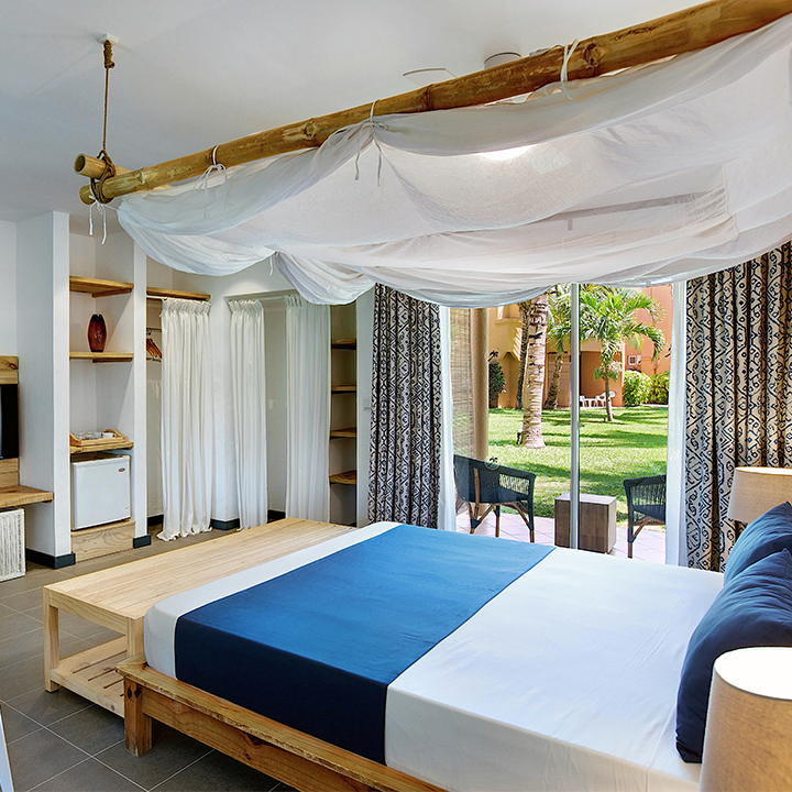 Spacious bedroom with wooden beam canopy and white draped fabric, blue and white bedding, and glass doors opening to a manicured courtyard with palm trees.