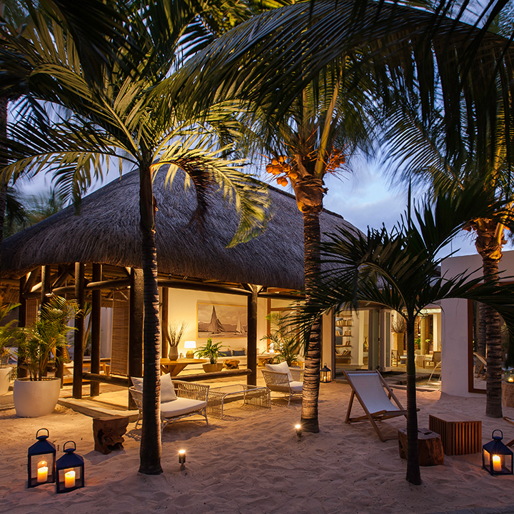Thatched-roof bungalow with illuminated interiors surrounded by palm trees, lounge chairs, and lanterns on sandy beach at dusk.