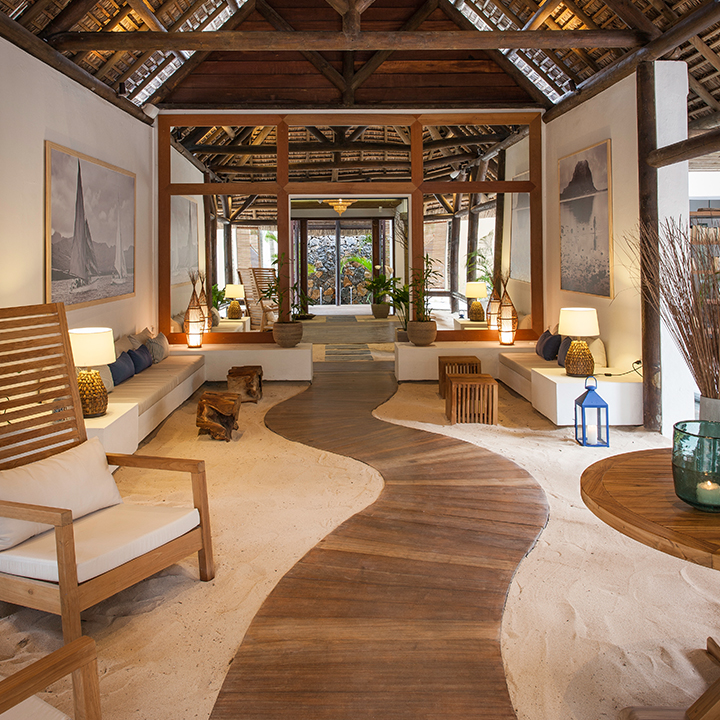 Open-air lobby with wooden beams, sand flooring, and winding wood boardwalk; wooden chairs, potted plants, and framed artwork line the space beneath a high thatched roof.