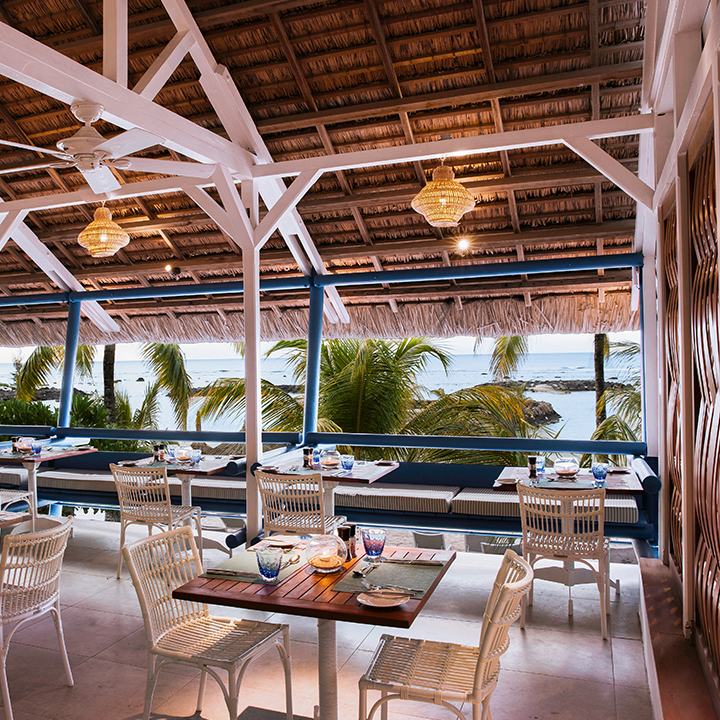 Beachfront dining pavilion at Veranda Pointe aux Biches with woven ceiling, white beams, wicker chairs, and ocean view beyond palm trees.